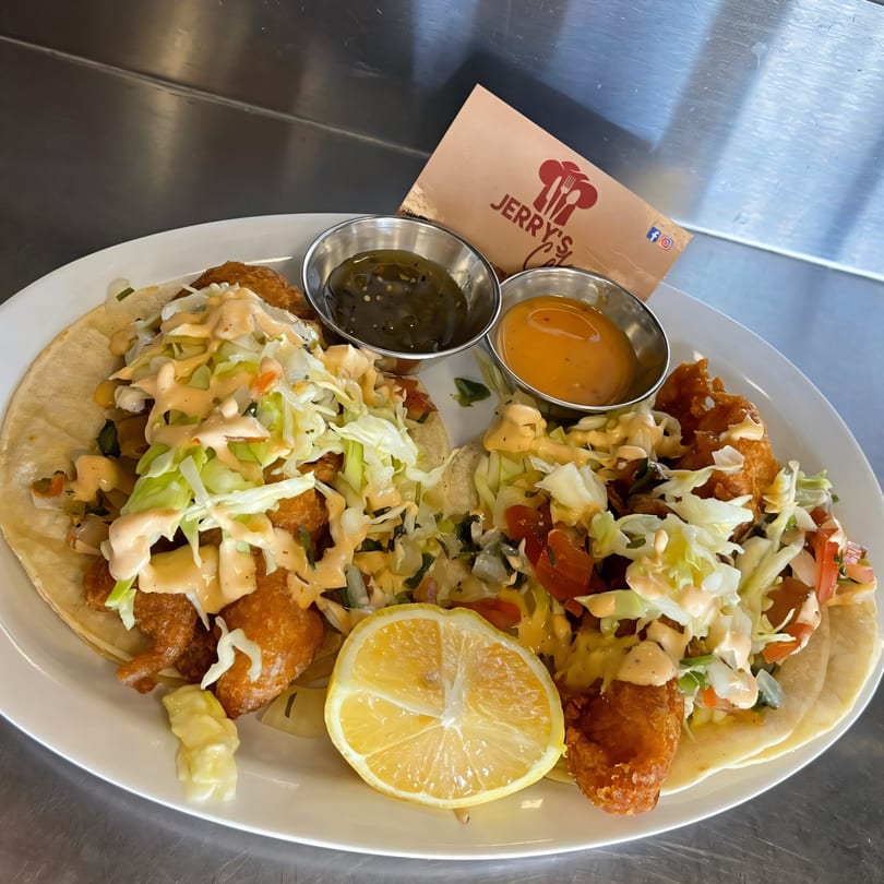 Two Jerry's Cafe fish tacos with shredded cabbage, sauce, lemon, and two dipping sauces.