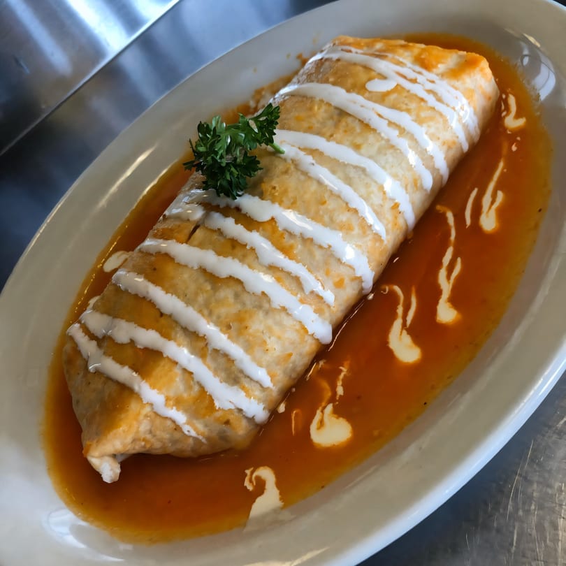 A large burrito covered in white sauce and parsley, resting in a red sauce on a white plate.