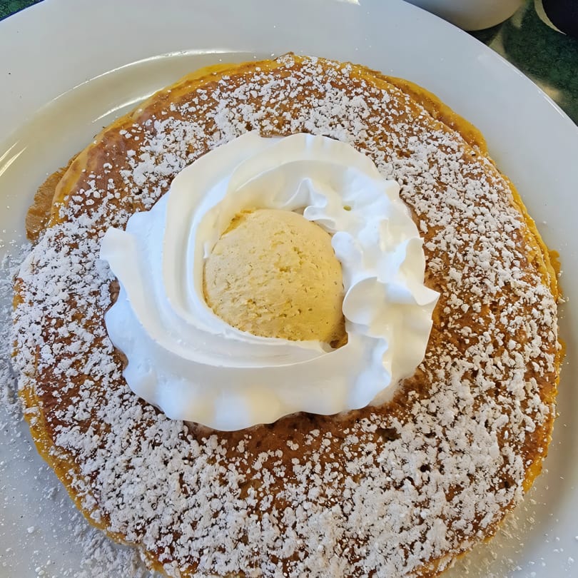 A large, round pancake covered in powdered sugar, topped with a scoop of ice cream and whipped cream.